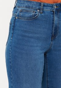 Augsta vidukļa zils denim stils džinsi ar gludu tekstūru, nelielu izbalēšanu, klasisku piecu kabatu dizainu un redzamām šuvēm.