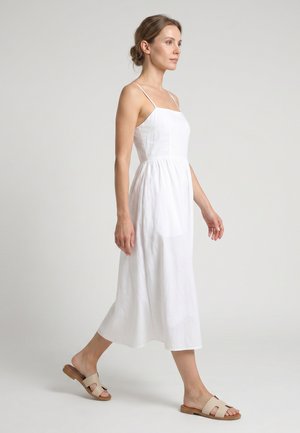 Vrouw loopt, gekleed in een witte mouwloze midi-jurk met dunne bandjes en beige platte slides op een effen lichte achtergrond.