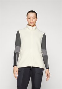 Beige Fleece-Weste mit hohem Kragen, Frontreißverschluss und seitlichen Taschen. Über einem dunklen Langarmoberteil getragen. Glatte Textur, minimalistisches Design.