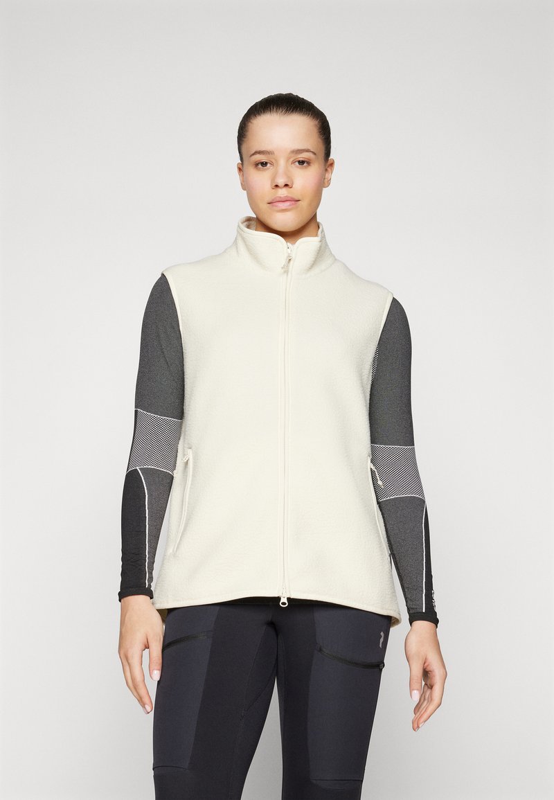 Beige Fleece-Weste mit hohem Kragen, Frontreißverschluss und seitlichen Taschen. Über einem dunklen Langarmoberteil getragen. Glatte Textur, minimalistisches Design.