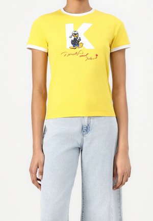 T-shirt jaune à manches courtes avec bordure blanche, avec un grand « K » blanc et un canard de dessin animé portant des lunettes de soleil, ainsi que le texte « Donald and Klan » en dessous.