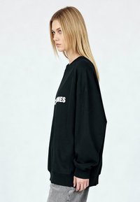 Sweat-shirt noir surdimensionné avec un col rond et des manches longues, présentant un graphisme texte blanc sur le devant et des détails de bord-côte à l'ourlet.