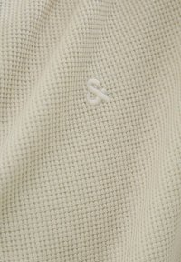 Tissu en crème texturé avec un motif à carreaux et un petit logo brodé dans le coin inférieur droit. Matière douce et légère.