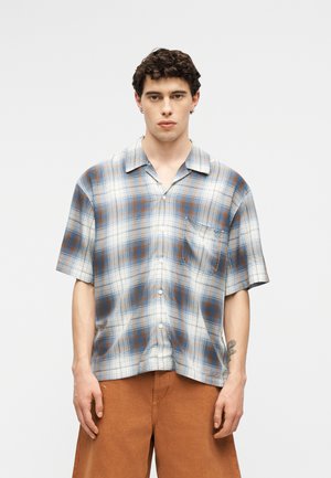 Mladić s kovrčavom kosom, nosi kratku kariranu košulju plave, bijele i smeđe boje s gumbima i smeđe šorceve, stoji ispred jednobojne pozadine.