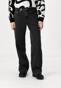 Zwarte wijde spijkerbroek van denim, met een hoge taille, vijf zakken en subtiele vervaging. Gecombineerd met zwarte sportschoenen.