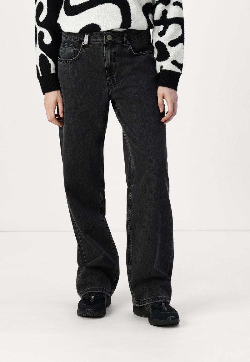 Zwarte wijde spijkerbroek van denim, met een hoge taille, vijf zakken en subtiele vervaging. Gecombineerd met zwarte sportschoenen.