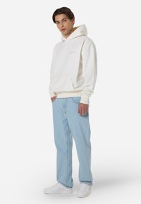 Λευκό φούτερ με κουκούλα και μπροστινή τσέπη, συνδυασμένο με ανοιχτό μπλε τζιν παντελόνι και λευκά αθλητικά παπούτσια. Άνετη εφαρμογή και απαλή υφή υφάσματος.