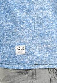 T-shirt bleu clair texturé avec un motif tacheté. Comprend une étiquette blanche avec du texte noir, un ourlet cousu et un tissu doux.