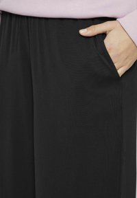 Crne, elastične hlače s mekim teksturom, s bočnim džepovima. Prikaz izbliza otkriva drapirane tkanine i detalje šava.