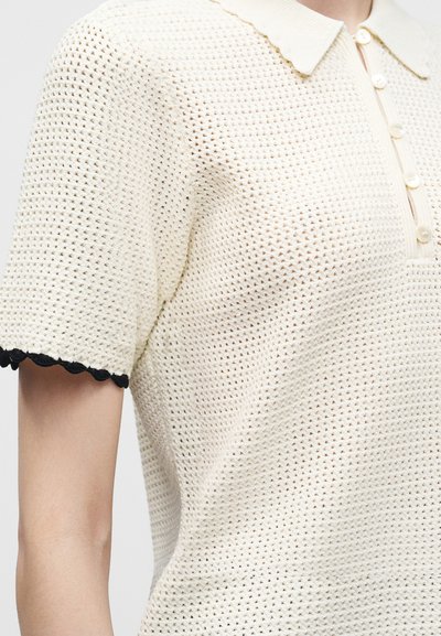 Gros plan d'un polo en maille crème à manches courtes avec col, patte de boutonnage et bords festonnés noirs aux manches.
