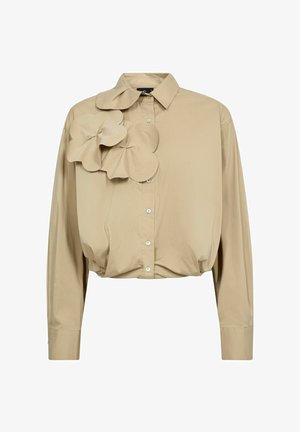 Cămașă bej scurtă, cu un accent mare și texturat în formă de floare pe partea stângă, având un guler clasic și deschidere cu nasturi în față.