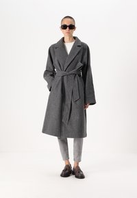 Sivi volneni plašč z pasom, prevelikimi dolgimi rokavi, stranskimi žepi in širokimi reverji, v kombinaciji s svetlimi kavbojkami in temnimi čevlji.