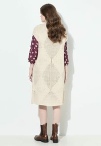 Beiges Strickkleid mit Rautenmuster, langen Ärmeln mit floralem Druck und seitlichem Schlitz. Getragen mit braunen Stiefeletten.