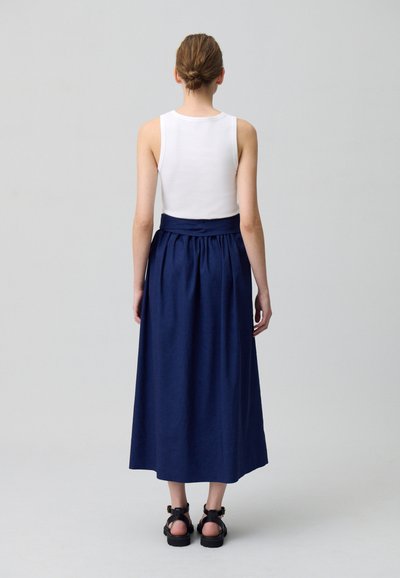 Jupe maxi bleu marine avec des fronces à la taille, associée à un débardeur blanc. Sandales plates visibles aux pieds, design minimaliste.