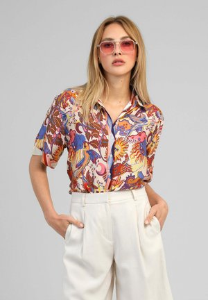 Femme portant une chemise colorée à manches courtes à motifs, un pantalon blanc taille haute et des lunettes de soleil teintées de rose, debout les mains dans les poches.