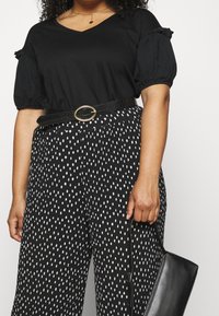 Černé tričko s výstřihem do V a nabíranými rukávy, doplněné širokými kalhotami s černobílým puntíkovaným vzorem, stažené černým páskem se zlatou sponou.