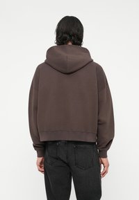 Sweat à capuche marron coupé, avec une grande capuche, fabriqué dans un tissu doux. Présente des coutures d'épaule tombantes et des poignets élastiques, associé à un jean foncé.