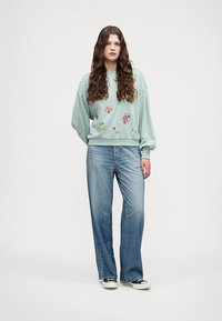 Menta zöld pulóver virágmintával, világoskék egyenes szabású farmerrel és fekete sportcipővel párosítva. Puha anyag, laza fazon.