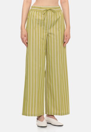 Donna che indossa pantaloni a gamba larga verde oliva con righe verticali bianche e scarpe basse bianche, in piedi davanti a uno sfondo semplice.