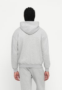 Braidelt hajú személy világosszürke kapucnis pulcsiban és azzal megegyező melegítőnadrágban áll, háttal a fényes fehér háttérnek.