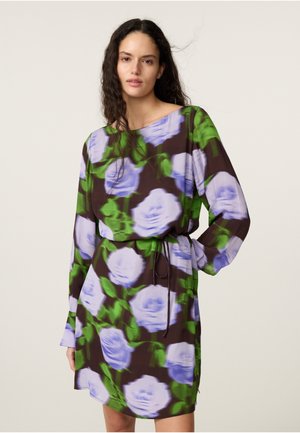 Vrouw met lang krullend haar die een los zittende bloemenjurk draagt met paarse bloemen en groene bladeren op een donkere achtergrond.