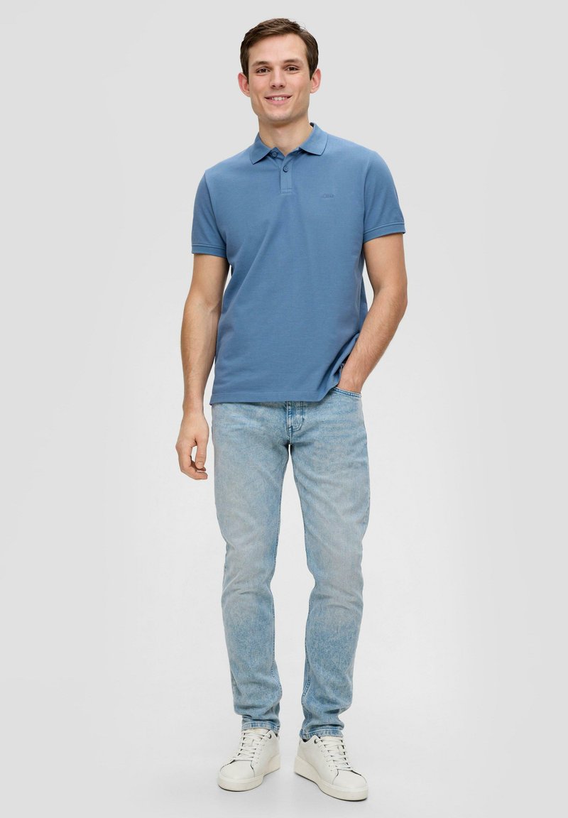 Polo shirt azzurro chiaro con colletto, abbinato a jeans chiari e sneakers bianche. Caratteristiche includono un logo discreto sul petto.