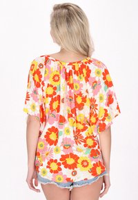 Blusa floreale con maniche corte a volant, caratterizzata da grandi fiori rossi, gialli e rosa su uno sfondo bianco. Dettaglio plissettato sul retro.