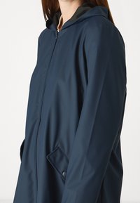 Marinblå vattentät jacka med dragkedja, huva och sidofickor; slät textur med en lätt glans och inga synliga mönster.