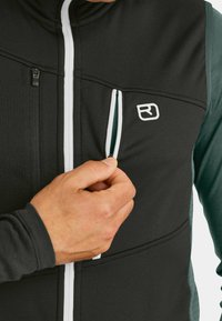 Schwarze und grüne zipbare Jacke mit einem weißen Akzent, strukturiertem Stoff und einer seitlichen Tasche mit Reißverschluss. Logo auf der Vorderseite sichtbar.