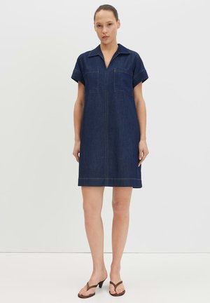 Vrouw staand, dragend een donker denimjurk met korte mouwen, kraag en voorzakken, gecombineerd met bruine sandalen met hakken.