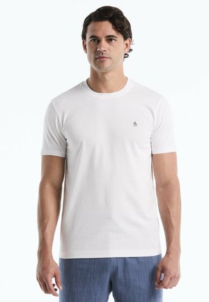 Ein Mann steht frontal und trägt ein schlichtes weißes T-Shirt mit kurzen Ärmeln, einem kleinen Pinguin-Logo und blaue strukturierte Hose.