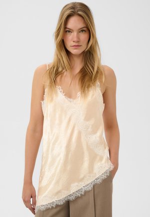 Jeune femme aux longs cheveux blonds portant un caraco en satin crème orné de dentelle et un pantalon taupe à jambes larges, debout devant un fond uni.