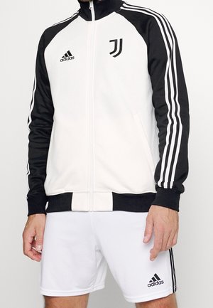 Homme portant une veste zippée Adidas Juventus noire et blanche avec un short Adidas blanc assorti.