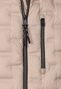 Beige vadderad jacka med en vertikal svart dragkedja, med kontrasterande texturerad sömnad och en dragsko med svarta detaljer.