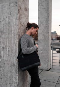 Black messenger bag with leather accents, hanging strap, and structured shape, next to a textured gray concrete pillar.