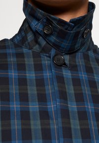 Giacca a quadri blu e verde con colletto alto e bottoni neri, indossata abbottonata da una persona, con il collo parzialmente visibile.