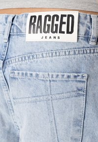 Jeans de mezclilla azul claro con una etiqueta de cuero blanca que dice "RAGGED JEANS". Las características incluyen un bolsillo trasero y detalles de costura visibles.