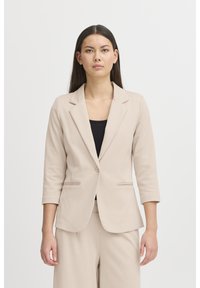 Frau mit langen dunklen Haaren, die einen beigen Blazer und passende Hosen über einem schwarzen Oberteil trägt, vor einem schlichten hellen Hintergrund stehend.