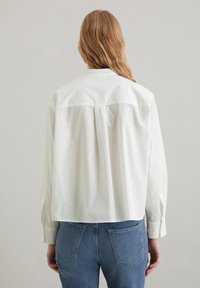 Valkoinen puuvillainen pitkähihainen paita, jossa on kaulus, lyhennetty malli ja laskos takana, yhdistetty sinisiin farkkuihin.