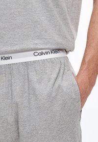 Hallid puuvillased pidžaamapüksid elastse vöökohaga, millel on valge riba ja must "Calvin Klein" logo. Esitasku nähtav.