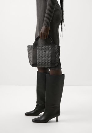 Kobieta ubrana w szarą, prążkowaną sukienkę i czarne kozaki do kolan, trzymająca po bokach szarą torbę na ramię w czarne wzory z czarnymi uchwytami.