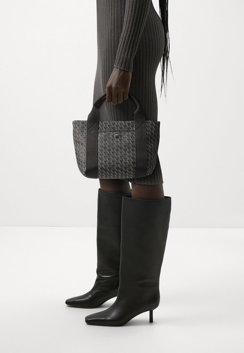 Femme portant une robe grise côtelée et des bottes noires montantes, tenant à ses côtés un sac fourre-tout gris à motif avec des poignées noires.