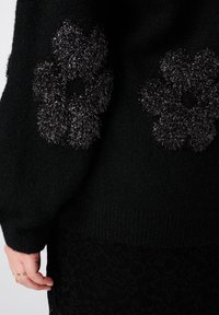 Pull noir à texture douce, orné de deux appliqués de fleurs duveteuses en gris foncé. Détail de poignets et d'ourlet côtelés.