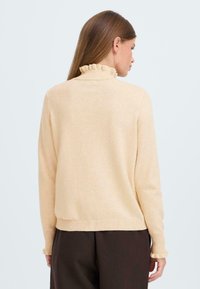 Pull beige clair avec un col haut et festonné, des manches longues et des poignets côtelés, associé à un pantalon marron foncé. Texture tricot lisse.