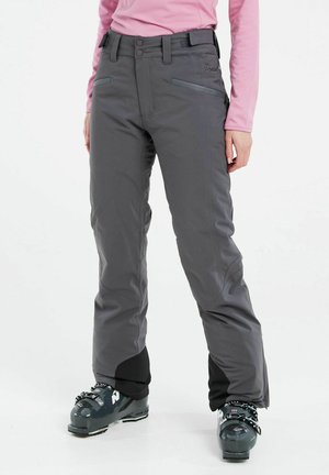 Femme portant un pantalon de ski gris, des chaussures de ski noires et un haut rose à manches longues, debout sur un fond blanc.