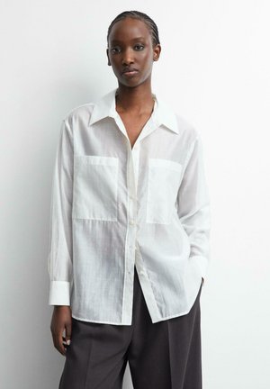 Vrouw met gevlochten haar die een losse witte blouse met knopen en donkere wijde broek draagt, staand tegen een witte achtergrond.