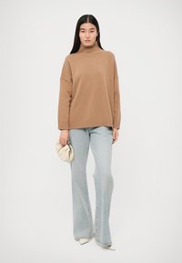 Maglione a collo alto beige oversize con orlo a coste, abbinato a jeans svasati blu chiaro. Tiene una pochette color crema. Sfondo pulito.