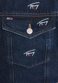 Denimjacka i mörkblått tyg med kontrasterande guldsömmar och broderade logotyper. Inkluderar metallknappar och bröstfickor.