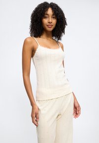 Jeune femme aux cheveux bouclés portant un caraco crème et un pantalon assorti avec petits motifs de cœurs roses sur fond blanc.