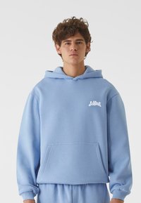 Šviesiai mėlynas džemperis su minkšta medžiaga, turintis priekinę kišenę ir baltą logotipą kairėje krūtinės pusėje. Įeina reguliuojama kapišoną.